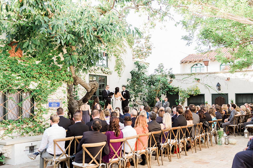 Fall wedding at the villa san juan capistrano bride strapless mermaid fit gown with a sweetheart neckline and ribbon belt with bow in the back with groom black notch lapel suit with a white dress shirt and black bow tie with a black and white plaid pocket square and white floral boutonniere holding hands during ceremony far away