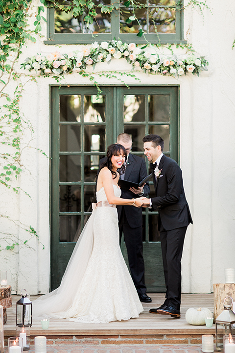 Fall wedding at the villa san juan capistrano bride strapless mermaid fit gown with a sweetheart neckline and ribbon belt with bow in the back with groom black notch lapel suit with a white dress shirt and black bow tie with a black and white plaid pocket square and white floral boutonniere holding hands during ceremony and laughing