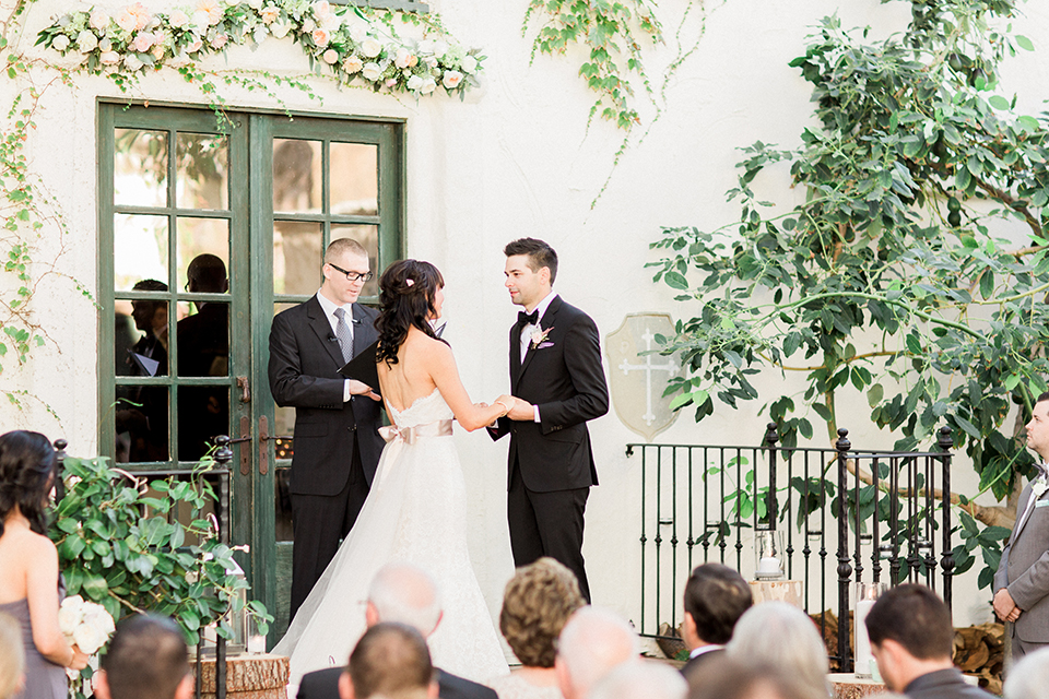 Fall wedding at the villa san juan capistrano bride strapless mermaid fit gown with a sweetheart neckline and ribbon belt with bow in the back with groom black notch lapel suit with a white dress shirt and black bow tie with a black and white plaid pocket square and white floral boutonniere holding hands during ceremony smiling