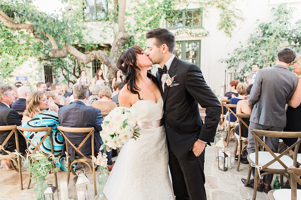 Fall wedding at the villa san juan capistrano bride strapless mermaid fit gown with a sweetheart neckline and ribbon belt with bow in the back with groom black notch lapel suit with a white dress shirt and black bow tie with a black and white plaid pocket square and white floral boutonniere kissing after ceremony walking back down the aisle