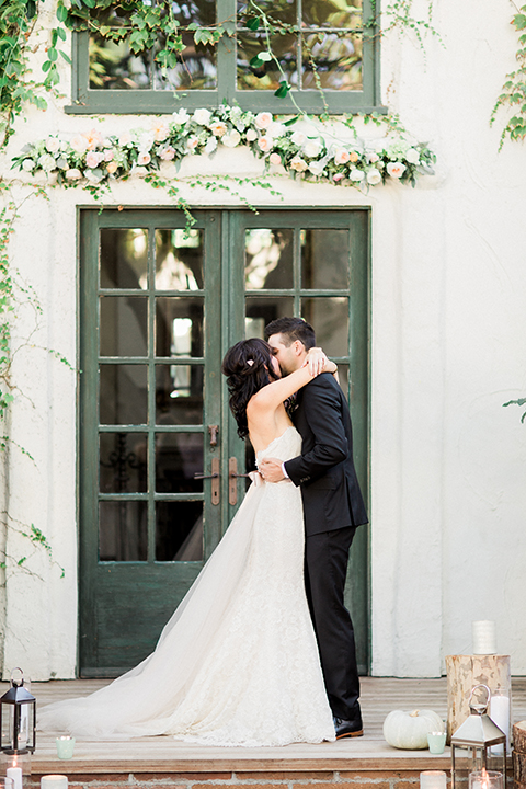 Fall wedding at the villa san juan capistrano bride strapless mermaid fit gown with a sweetheart neckline and ribbon belt with bow in the back with groom black notch lapel suit with a white dress shirt and black bow tie with a black and white plaid pocket square and white floral boutonniere kissing during ceremony