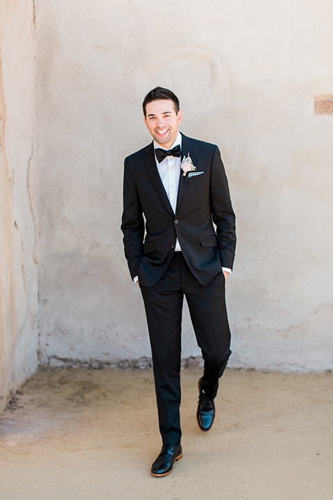 Fall wedding at the villa san juan capistrano groom black notch lapel suit with a white dress shirt and black bow tie with a black and white plaid pocket square with a white floral boutonniere smiling with hands in pockets walking