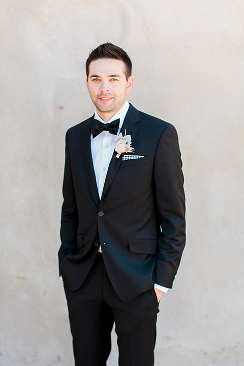 Fall wedding at the villa san juan capistrano groom black notch lapel suit with a white dress shirt and black bow tie with a black and white plaid pocket square with a white floral boutonniere smiling with hands in pockets
