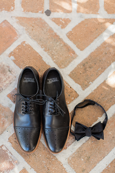 Fall wedding at the villa san juan capistrano groom black notch lapel suit with a white dress shirt and black bow tie with a black and white plaid pocket square with a white floral boutonniere smiling with hands in pockets with black shoes and black bow tie on brick ground for photo