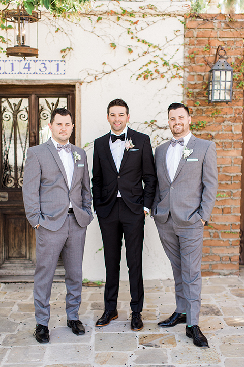 Fall wedding at the villa san juan capistrano groom black notch lapel suit with a white dress shirt and black bow tie with a black and white plaid pocket square with a white floral boutonniere smiling with hands in pockets standing with groomsmen grey notch lapel suits with matching grey striped bow ties and white floral boutonnieres