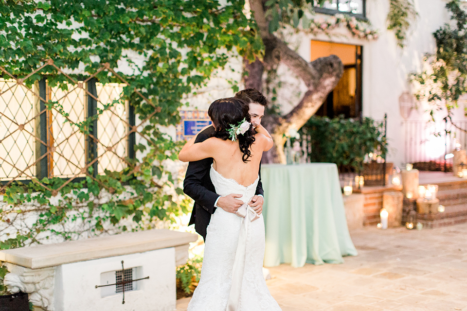 Fall wedding at the villa san juan capistrano bride strapless mermaid fit gown with a sweetheart neckline and ribbon belt with bow in the back with groom black notch lapel suit with a white dress shirt and black bow tie with a black and white plaid pocket square and white floral boutonniere hugging at reception first dance