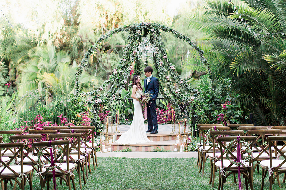 Los angeles outdoor wedding at eden gardens bride form fitting gown with draping detail and open back design with thin straps and groom navy blue shawl lapel tuxedo with a matching vest and white dress shirt with a black and white gingham plaid bow tie and pink floral boutonniere standing during ceremony by floral altar hugging and bride holding green and pink floral bridal bouquet with ribbon decor far away