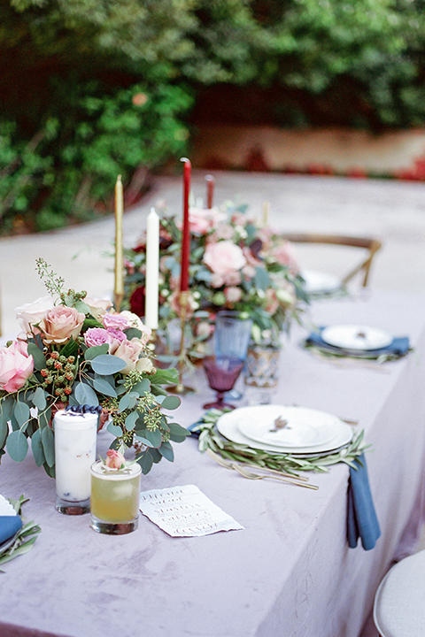 Los angeles outdoor wedding at eden gardens table set up with light grey table linen and white place setting with greenery and pink flower centerpiece decor and blue linen napkin with tall white and red candle decor and brown wood rustic chairs