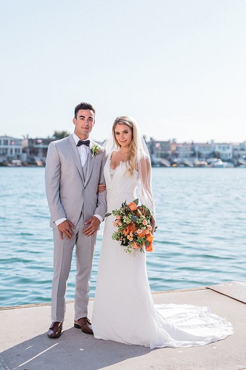 Newport beach wedding at balboa bay resort bride form fitting lace gown with short lace sleeves and a plunging neckline with lace decor with groom light grey peak lapel suit with matching vest and white dress shirt with a charcoal grey bow tie and green floral boutonniere standing holding hands and bride holding green colorful floral bouquet