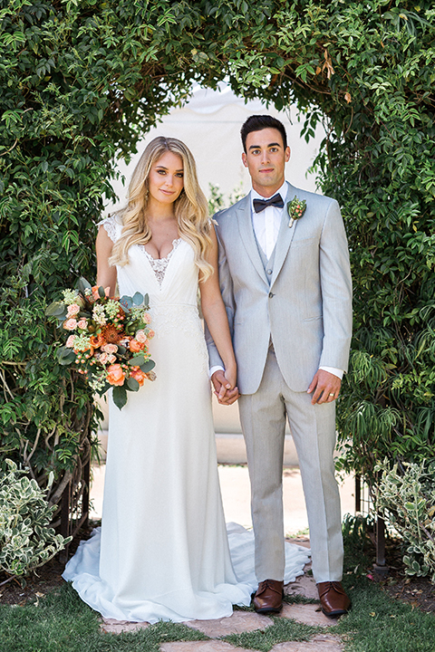 Newport beach wedding at balboa bay resort bride form fitting lace gown with short lace sleeves and a plunging neckline with lace decor with groom light grey peak lapel suit with matching vest and white dress shirt with a charcoal grey bow tie and green floral boutonniere standing holding hands and bride holding green colorful floral bouquet