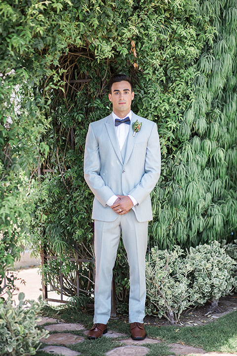 Newport beach wedding at balboa bay resort groom light grey peak lapel suit with a matching vest and white dress shirt with a charcoal grey bow tie and green floral boutonniere standing with hands folded