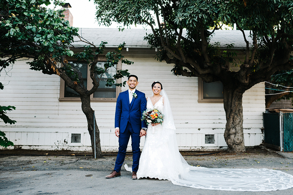 San diego autumn inspired outdoor wedding at limoneira ranch bride form fitting lace gown with straps and a v neckline with open back design and long veil with groom cobalt notch lapel suit by allure men with a matching vest and white dress shirt with a long white tie and white floral boutonniere holding hands and bride holding red and green floral bridal bouquet