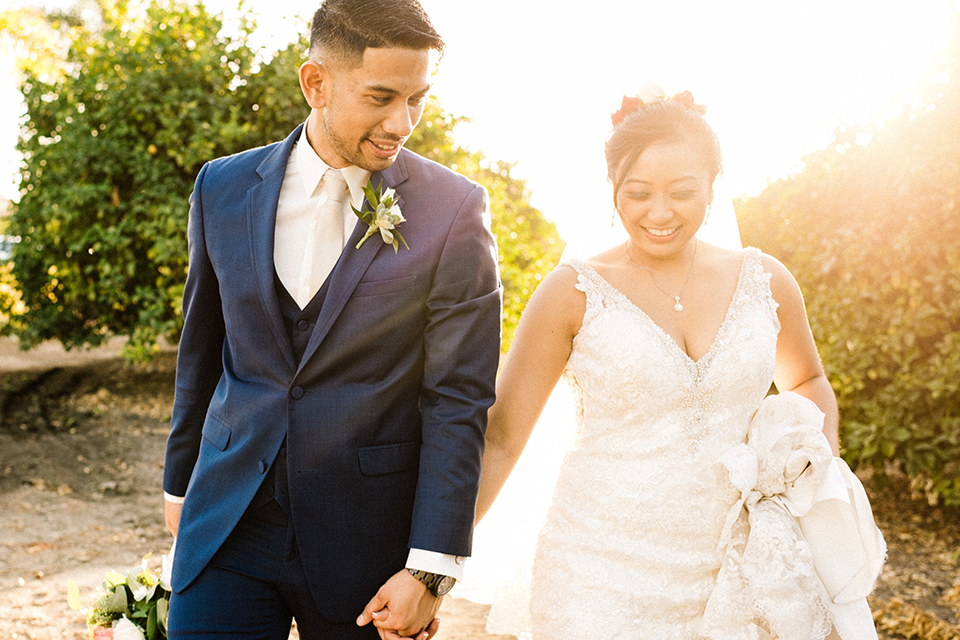 San diego autumn inspired outdoor wedding at limoneira ranch bride form fitting lace gown with straps and a v neckline with open back design and long veil with groom cobalt notch lapel suit by allure men with a matching vest and white dress shirt with a long white tie and white floral boutonniere walking and holding hands at sunset