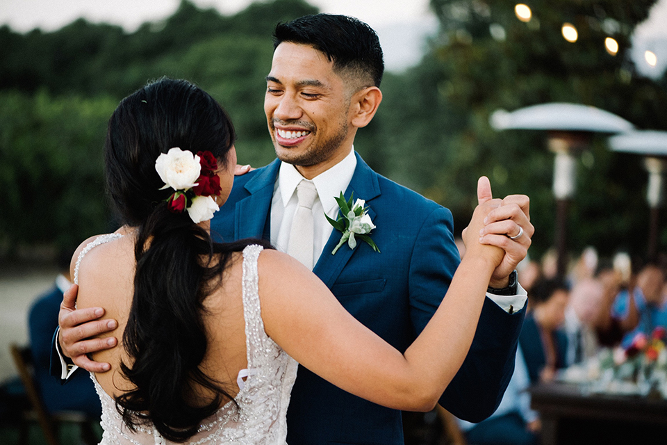 San diego autumn inspired outdoor wedding at limoneira ranch bride form fitting lace gown with straps and a v neckline with open back design and long veil with groom cobalt notch lapel suit by allure men with a matching vest and white dress shirt with a long white tie and white floral boutonniere holding hands and dancing close up