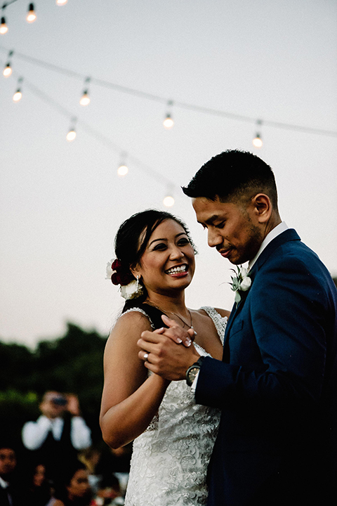 San diego autumn inspired outdoor wedding at limoneira ranch bride form fitting lace gown with straps and a v neckline with open back design and long veil with groom cobalt notch lapel suit by allure men with a matching vest and white dress shirt with a long white tie and white floral boutonniere holding hands and dancing