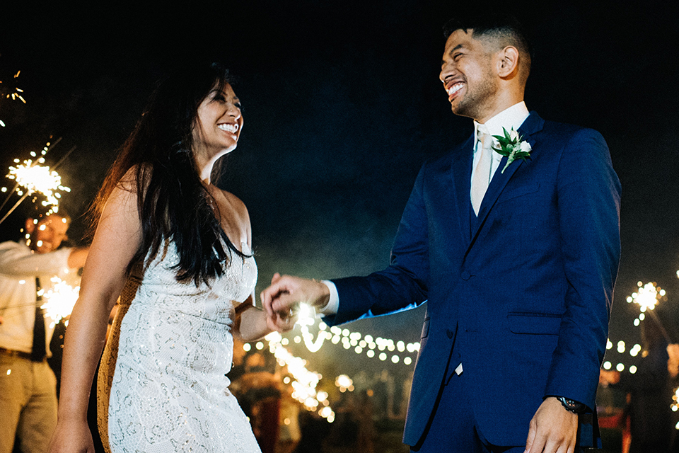 San diego autumn inspired outdoor wedding at limoneira ranch bride form fitting lace gown with straps and a v neckline with open back design and long veil with groom cobalt notch lapel suit by allure men with a matching vest and white dress shirt with a long white tie and white floral boutonniere holding hands and smiling sparkler exit