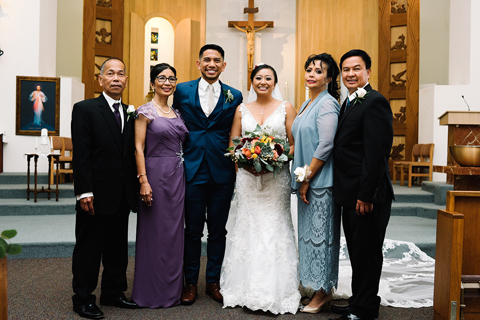 San diego autumn inspired outdoor wedding at limoneira ranch bride form fitting lace gown with straps and a v neckline with open back design and long veil with groom cobalt notch lapel suit by allure men with a matching vest and white dress shirt with a long white tie and white floral boutonniere standing with parents 