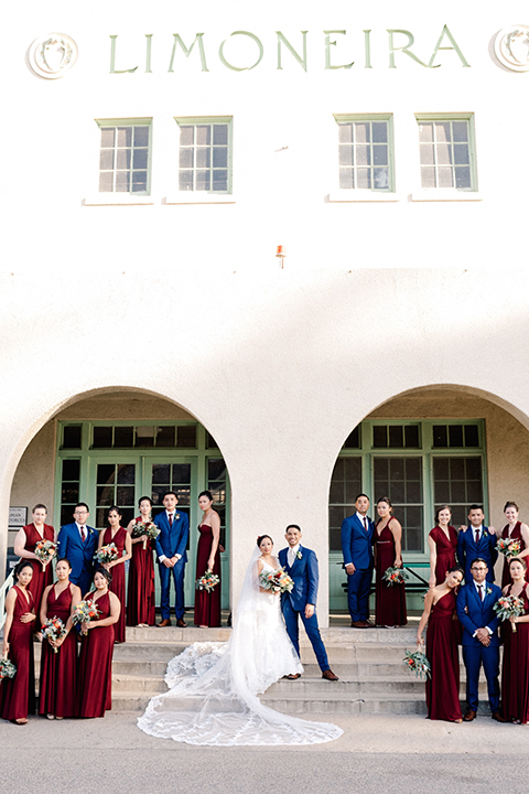 San diego autumn inspired outdoor wedding at limoneira ranch bride form fitting lace gown with straps and a v neckline with open back design and long veil with groom cobalt notch lapel suit by allure men with a matching vest and white dress shirt with a long white tie and white floral boutonniere standing with wedding party bridesmaids long burgundy dresses and groomsmen cobalt blue suits with long burgundy ties