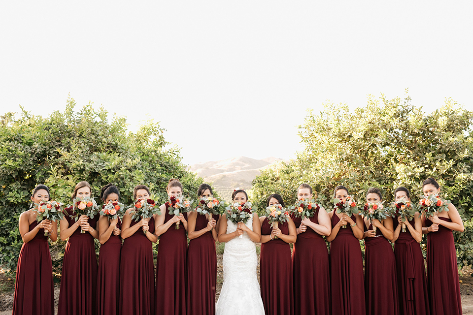 San diego autumn inspired outdoor wedding at limoneira ranch bride form fitting lace gown with straps and a v neckline with open back design and long veil holding green and dark red floral bridal bouquet standing with bridesmaids long burgundy dresses holding floral bouquets close to face