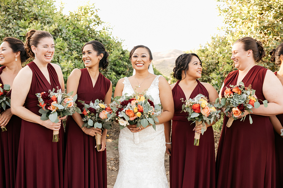 San diego autumn inspired outdoor wedding at limoneira ranch bride form fitting lace gown with straps and a v neckline with open back design and long veil holding green and dark red floral bridal bouquet standing with bridesmaids long burgundy dresses holding floral bouquets and smiling