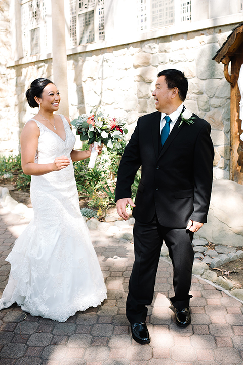 San diego autumn inspired outdoor wedding at limoneira ranch bride form fitting lace gown with straps and a v neckline with open back design and long veil holding green and dark red floral bridal bouquet standing with dad after first look navy blue suit with long cobalt blue tie smiling