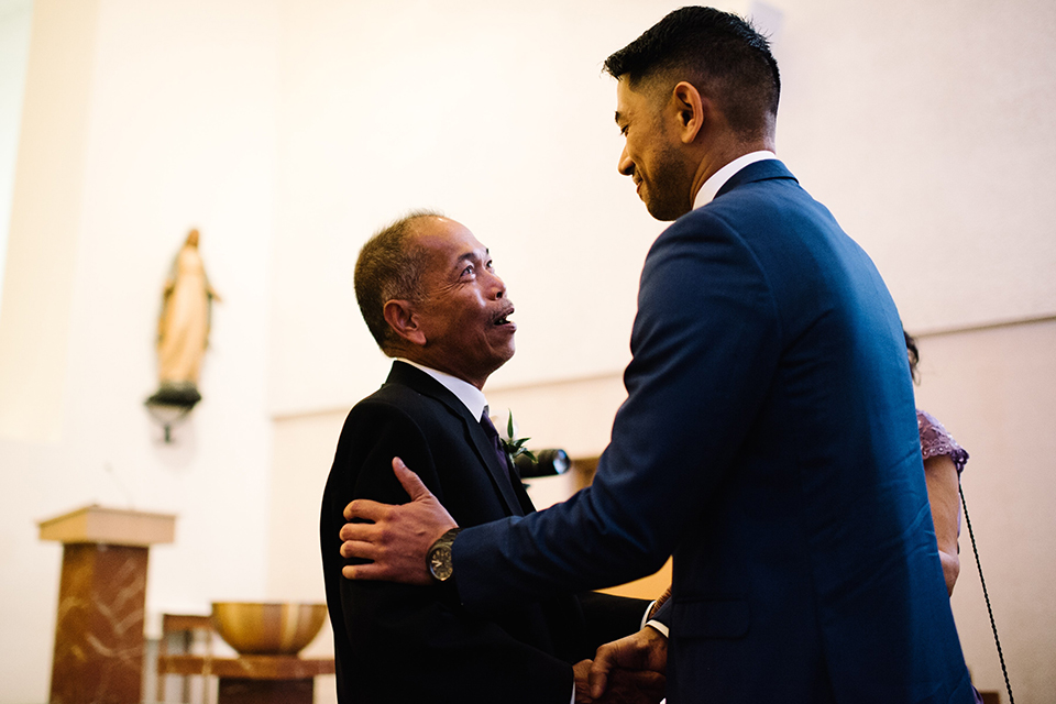 San diego autumn inspired outdoor wedding at limoneira ranch groom cobalt blue notch lapel suit with a matching vest and white dress shirt with a long white tie and white floral boutonniere shaking hands with dad