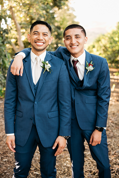 San diego autumn inspired outdoor wedding at limoneira ranch groom cobalt blue notch lapel suit with a matching vest and white dress shirt with a long white tie and white floral boutonniere standing with groomsman cobalt blue suit with long burgundy tie and burgundy floral boutonniere