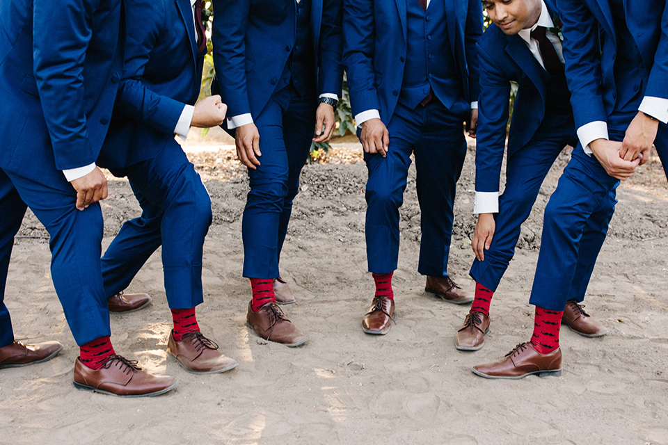 San diego autumn inspired outdoor wedding at limoneira ranch groom cobalt blue notch lapel suit with a matching vest and white dress shirt with a long white tie and white floral boutonniere standing with groomsmen cobalt blue suits close up on brown shoes with red mustache socks fun wedding photo idea for groomsmen