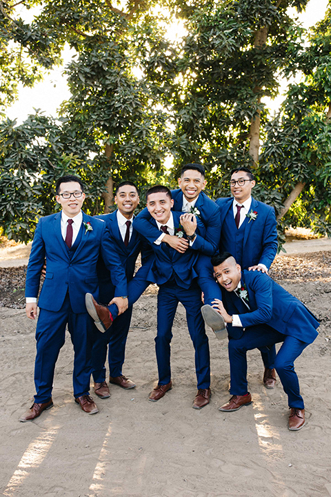 San diego autumn inspired outdoor wedding at limoneira ranch groom cobalt blue notch lapel suit with a matching vest and white dress shirt with a long white tie and white floral boutonniere standing with groomsmen cobalt blue suits with long burgundy ties and burgundy floral boutonnieres smiling