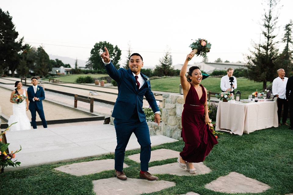 San diego autumn inspired outdoor wedding at limoneira ranch reception wedding party entrance bridesmaid long burgundy dress with red and green floral bouquet walking in with groomsman cobalt blue suit with a long burgundy tie and burgundy floral boutonniere