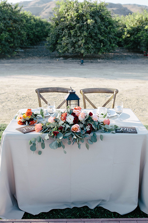 San diego autumn inspired outdoor wedding at limoneira ranch table set up light blue table linen with light orange and green flower centerpiece decor with light brown wooden rustic chairs and lantern and candle decor