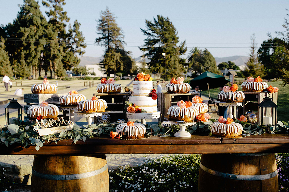 San diego autumn inspired outdoor wedding at limoneira ranch table set up light blue table linen with light orange and green flower centerpiece decor with light brown wooden rustic chairs and lantern and candle decor with wood dessert table and assorted cakes and dessert