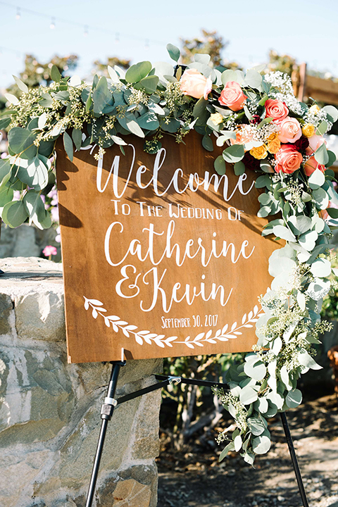 San diego autumn inspired outdoor wedding at limoneira ranch wedding welcome sign light brown wood rustic sign with white calligraphy writing for bride and groom with greenery floral decor on top standing on top of easel wedding photo idea for welcome sign