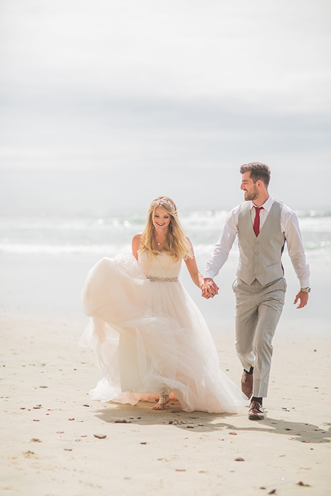 San diego beach wedding at blacks beach bride strapless ball gown with a sweetheart neckline and crystal belt and headband with groom light grey peak lapel suit by ike behar with a matching vest and white dress shirt with a long red matte tie and red flower lapel pin running and holding hands