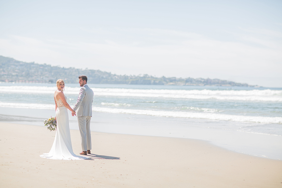 San diego beach wedding at blacks beach bride strapless ball gown with a sweetheart neckline and crystal belt and headband with groom light grey peak lapel suit by ike behar with a matching vest and white dress shirt with a long red matte tie and red flower lapel pin standing on the beach far away holding hands