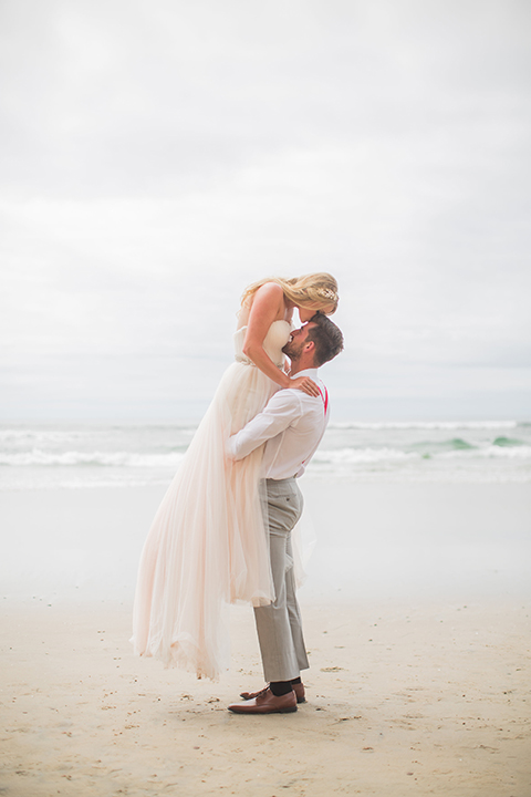 San diego beach wedding at blacks beach bride strapless ball gown with a sweetheart neckline and crystal belt and headband with groom light grey peak lapel suit by ike behar with a matching vest and white dress shirt with a long red matte tie and red flower lapel pin groom lifting bride touching heads and hugging