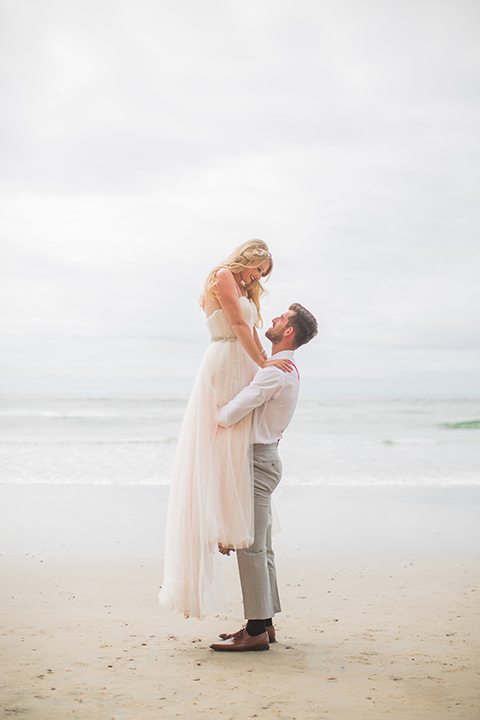 San diego beach wedding at blacks beach bride strapless ball gown with a sweetheart neckline and crystal belt and headband with groom light grey peak lapel suit by ike behar with a matching vest and white dress shirt with a long red matte tie and red flower lapel pin groom lifting bride smiling