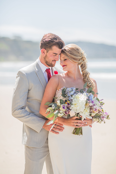 San diego beach wedding at blacks beach bride strapless ball gown with a sweetheart neckline and crystal belt and headband with groom light grey peak lapel suit by ike behar with a matching vest and white dress shirt with a long red matte tie and red flower lapel pin hugging and bride holding pink and blue floral bridal bouquet