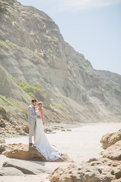 San diego beach wedding at blacks beach bride strapless ball gown with a sweetheart neckline and crystal belt and headband with groom light grey peak lapel suit by ike behar with a matching vest and white dress shirt with a long red matte tie and red flower lapel pin standing on the rocks on the beach hugging far away