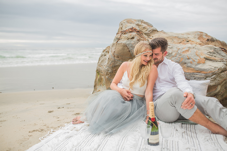 San diego beach wedding at blacks beach bride white top with light dusty blue tulle skirt and headband with groom light grey suit by ike behar with a white dress shirt and no tie sitting on blanket on the beach with bottle of champagne hugging