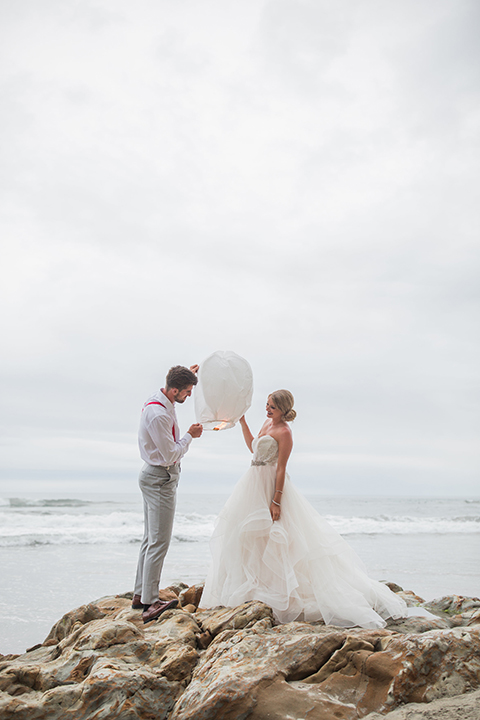 San diego beach wedding at blacks beach bride strapless ball gown with a sweetheart neckline and crystal belt and headband with groom light grey peak lapel suit by ike behar with a matching vest and white dress shirt with a long red matte tie and red flower lapel pin standing on the rocks holding white lantern