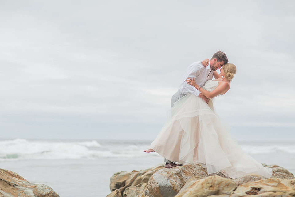 San diego beach wedding at blacks beach bride strapless ball gown with a sweetheart neckline and crystal belt and headband with groom light grey peak lapel suit by ike behar with a matching vest and white dress shirt with a long red matte tie and red flower lapel pin standing on the rocks hugging and smiling