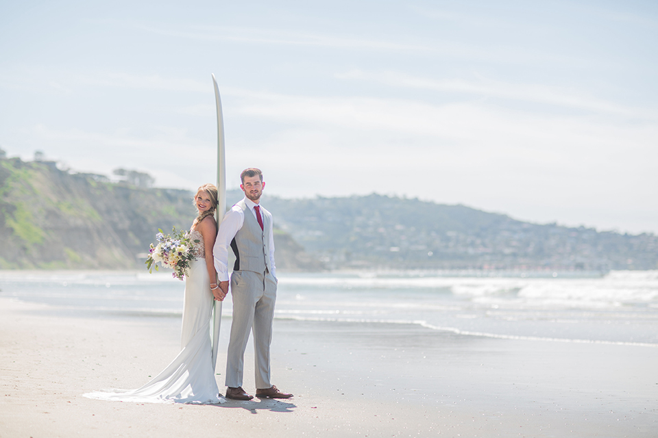 San diego beach wedding at blacks beach bride strapless ball gown with a sweetheart neckline and crystal belt and headband with groom light grey peak lapel suit by ike behar with a matching vest and white dress shirt with a long red matte tie and red flower lapel pin standing on the beach by surfboard holding hands