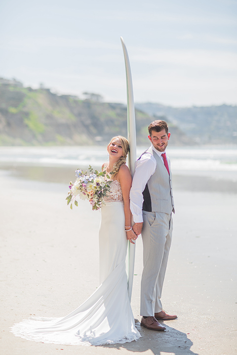 San diego beach wedding at blacks beach bride strapless ball gown with a sweetheart neckline and crystal belt and headband with groom light grey peak lapel suit by ike behar with a matching vest and white dress shirt with a long red matte tie and red flower lapel pin standing on the beach with surfboard holding hands and smiling