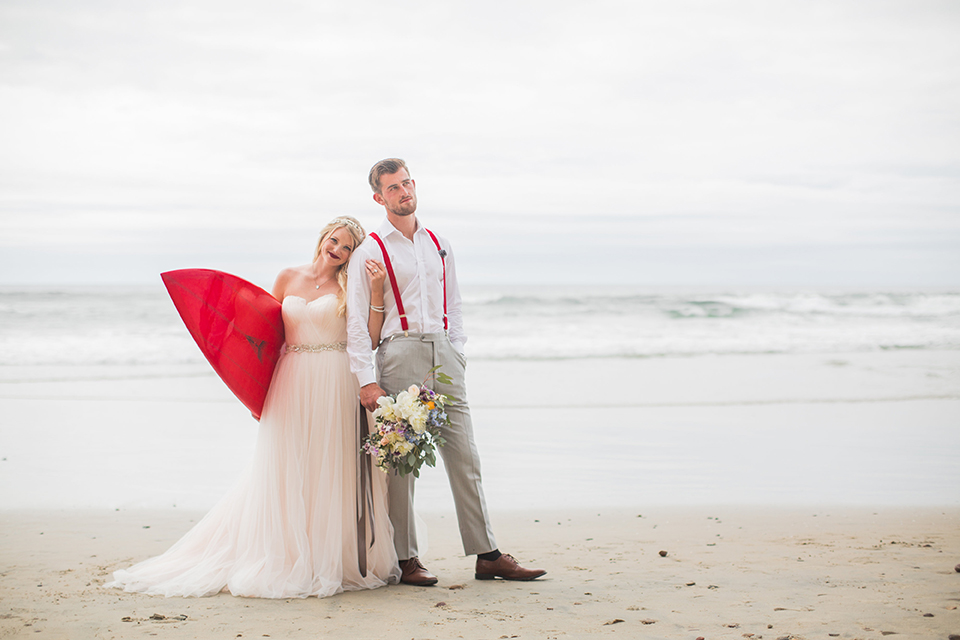 San diego beach wedding at blacks beach bride strapless ball gown with a sweetheart neckline and crystal belt and headband with groom light grey peak lapel suit by ike behar with a matching vest and white dress shirt with a long red matte tie and red flower lapel pin standing on the beach bride holding red surfboard and groom holding pink and blue floral bridal bouquet for bride