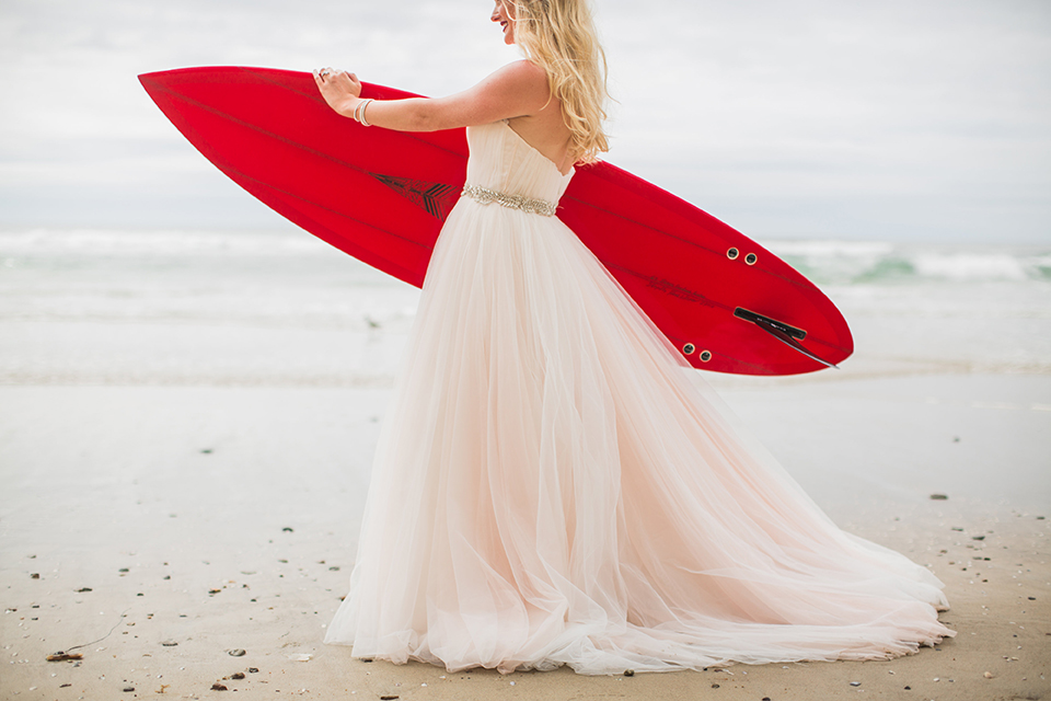 San diego beach wedding at blacks beach bride strapless ball gown with a sweetheart neckline and crystal belt and headband holding red surfboard under arm standing on the beach close up