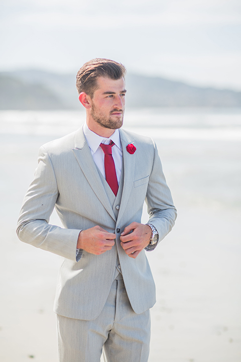 San diego beach wedding at blacks beach groom light grey peak lapel suit by ike behar with a matching vest and white dress shirt with a long red matte tie and matching red flower lapel pin buttoning coat in the front