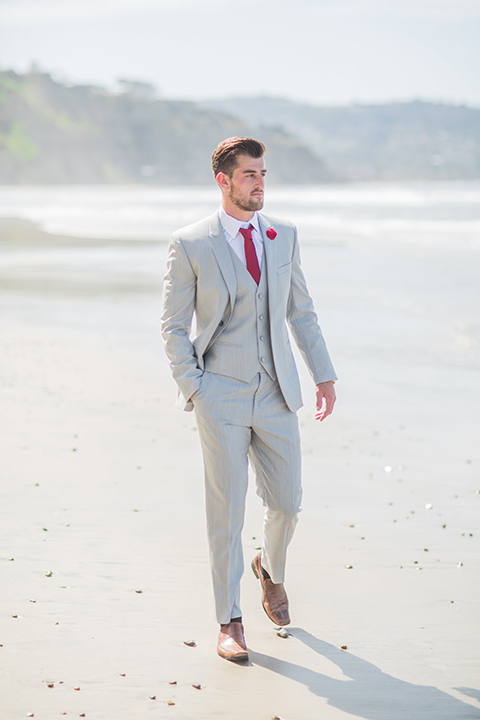 San diego beach wedding at blacks beach groom light grey peak lapel suit by ike behar with a matching vest and white dress shirt with a long red matte tie and matching red flower lapel pin walking on the beach with hand in pocket