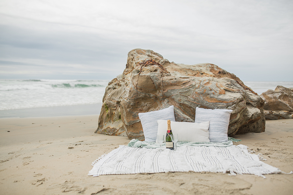 San diego beach wedding at blacks beach white blanket set up on the sand with rocks behind and light blue decorative pillows with light blue and pink flower decor and crystal wine glasses with blue geode rock name plates for bride and groom in calligraphy writing wedding photo idea for beach blanket set up
