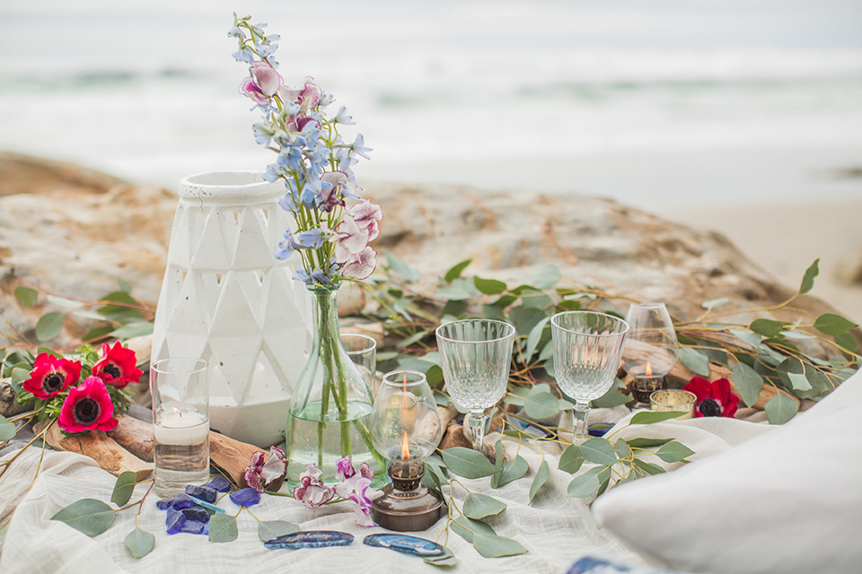 San diego beach wedding at blacks beach white blanket set up on the sand with rocks behind and light blue decorative pillows with light blue and pink flower decor and crystal wine glasses with blue geode rock name plates for bride and groom in calligraphy writing wedding photo idea for beach blanket set up